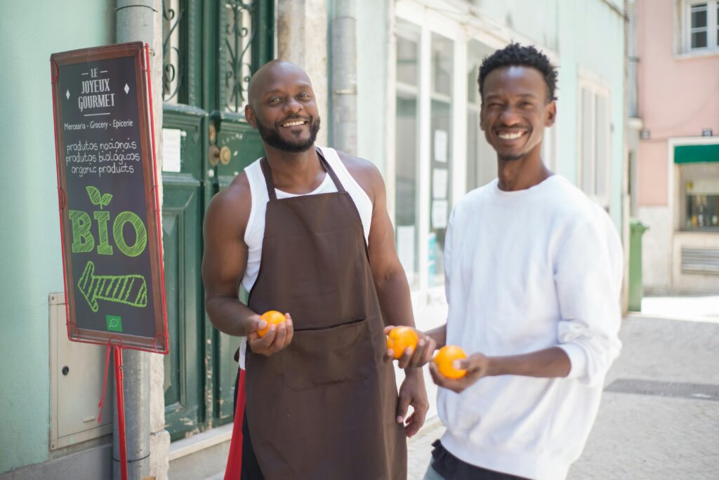 Two happy men in Lisbon holding oranges in front of a bio grocery store, promoting organic produce.
